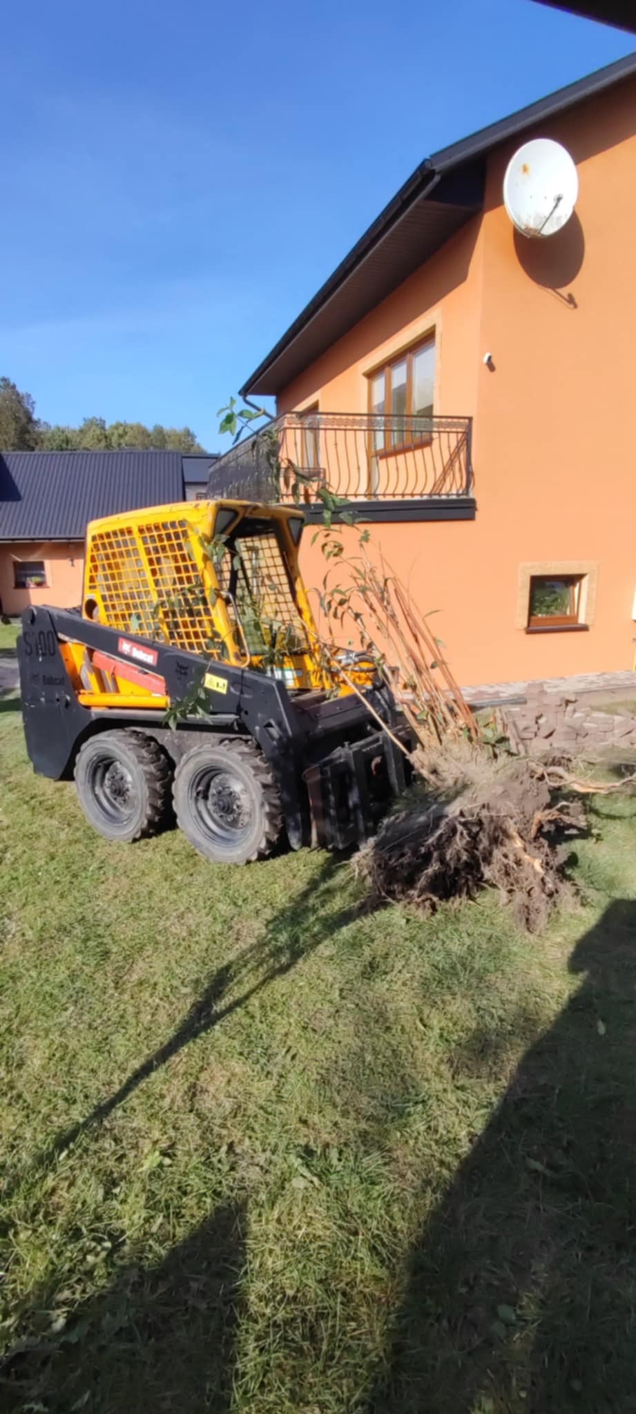Żółto-czarna miniładowarka Bobcat podnosi karpę drzewa na trawniku przed domem z pomarańczową elewacją i balkonem z kutą balustradą w słoneczny dzień.