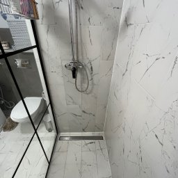 Oleksandr Vodopian - Wykończona łazienka z kabiną typu walk-in, wyłożona białymi płytkami imitującymi marmur. Widoczny odpływ liniowy, bateria prysznicowa i toaleta w odbiciu szyby.