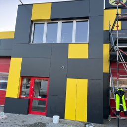 Oleksandr Vodopian - Elewacja budynku po ociepleniu, z szaro-żółtymi panelami i czerwoną stolarką. Widoczne rusztowania i narzędzia, sugerujące prace wykończeniowe.