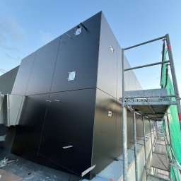 Oleksandr Vodopian - Czarna elewacja budynku w trakcie montażu, widoczna z rusztowania. Elementy mocujące na ścianie, błękitne niebo w tle. Ujęcie z dołu.