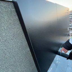 Oleksandr Vodopian - Fragment elewacji budynku z zamontowaną, czarną, metalową obróbką blacharską. Widoczna faktura elewacji i fragment dłoni w rękawie ochronnym.