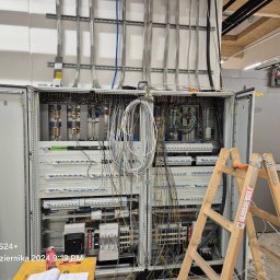Elektro-Instal Dawid Walden - Rozbudowana szafa elektryczna z otwartymi drzwiami, widoczne liczne przewody i elementy instalacji. Drabina oparta o szafę, sugerująca trwające prace instalacyjne.