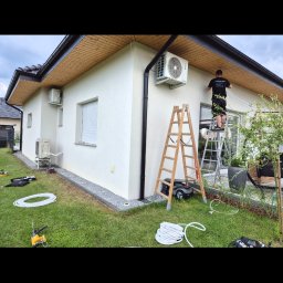 Elektro-Instal Dawid Walden - Technik na drabinie instaluje zewnętrzną jednostkę chłodzącą na białej ścianie domu. Obok leżą narzędzia i zwinięte rury, a w tle widać drugie urządzenie.