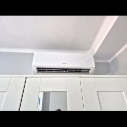 Elektro-Instal Dawid Walden - Biały klimatyzator ścienny Sinclair z cyfrowym wyświetlaczem '16', zamontowany wysoko na ścianie nad białymi szafkami z lustrzanymi panelami. Widoczny fragment sufitu z ozdobną listwą.