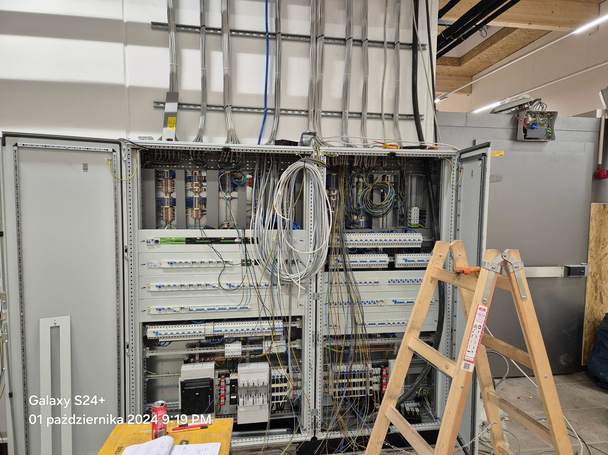 Rozbudowana szafa elektryczna z otwartymi drzwiami, widoczne liczne przewody i elementy instalacji. Drabina oparta o szafę, sugerująca trwające prace instalacyjne.