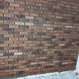 Marcin Urbanowski - Ściana wykończona cegłą klinkierową w odcieniach brązu i czerni, widoczne gniazdko elektryczne z wystającymi przewodami, dolna krawędź ściany w trakcie wykańczania.