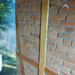 Ceglana ściana z drewnianymi elementami konstrukcyjnymi i tapetą z motywem leśnym w tle, widoczne przewody elektryczne i fragment instalacji wodnej.