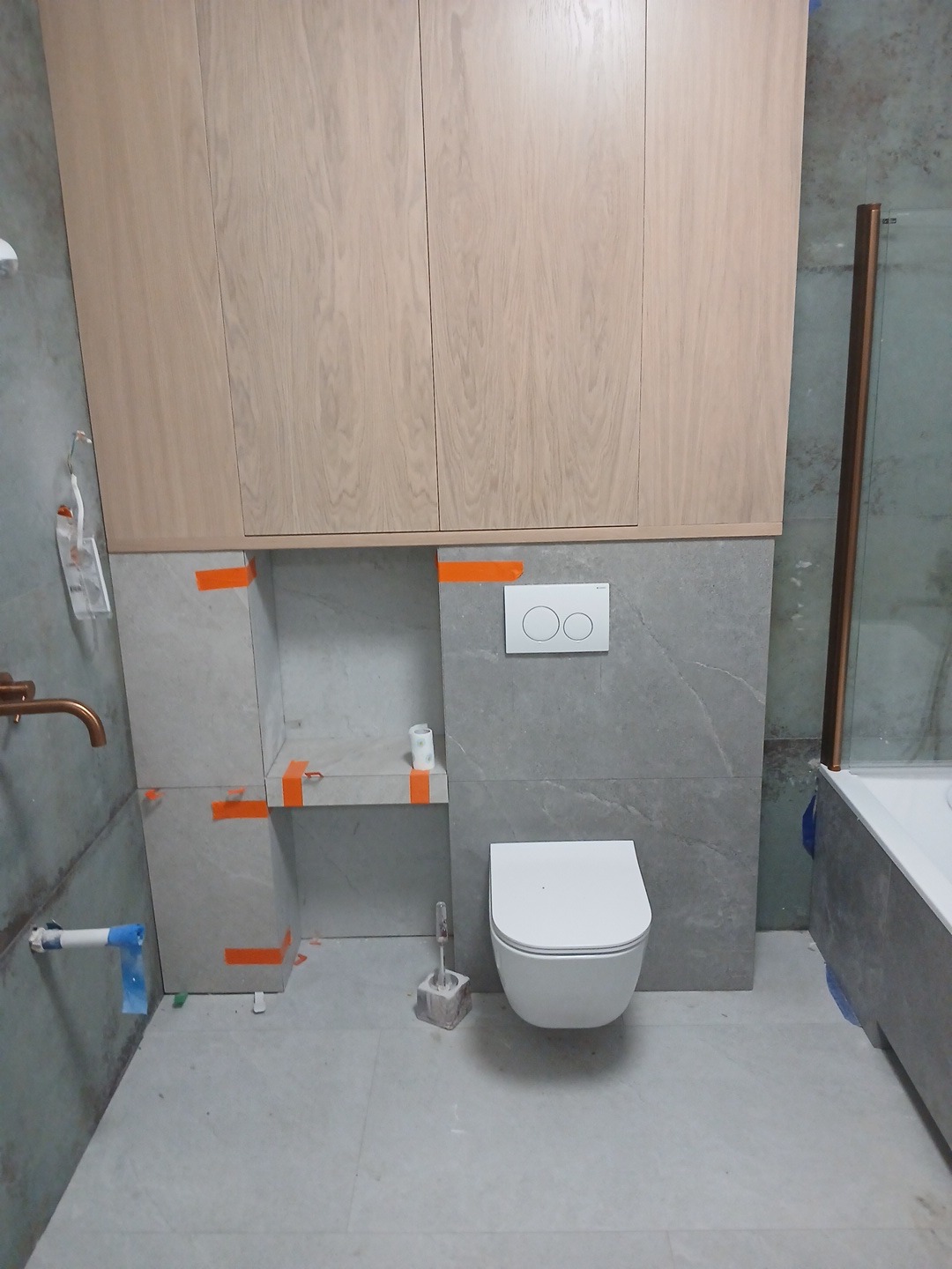 Nowoczesna łazienka w trakcie wykańczania: zabudowa szafkowa z jasnego drewna nad szarą ścianą z marmurowym wzorem, podwieszana toaleta, półka w zabudowie, widoczne taśmy malarskie, bateria...