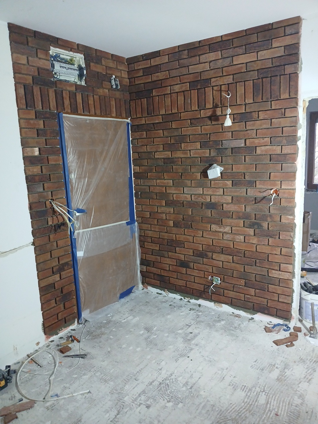 Montaż dekoracyjnej ściany z cegły klinkierowej w trakcie remontu mieszkania, zabezpieczone drzwi folią, widoczna instalacja elektryczna i narzędzia na podłodze.