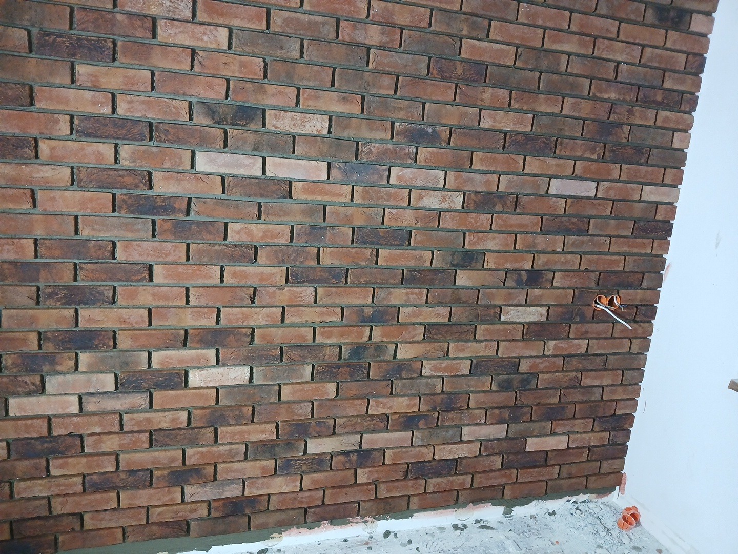 Ściana wykończona cegłą klinkierową w odcieniach brązu i czerni, widoczne gniazdko elektryczne z wystającymi przewodami, dolna krawędź ściany w trakcie wykańczania.