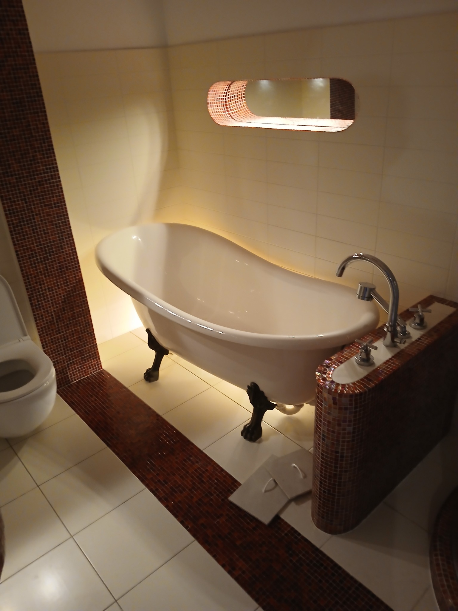 Biała wanna wolnostojąca na lwich nóżkach w łazience wyłożonej jasnymi płytkami, z dekoracyjnym pasem mozaiki w odcieniach brązu i podobną mozaiką na obudowie baterii, oświetlona od spodu i od góry...