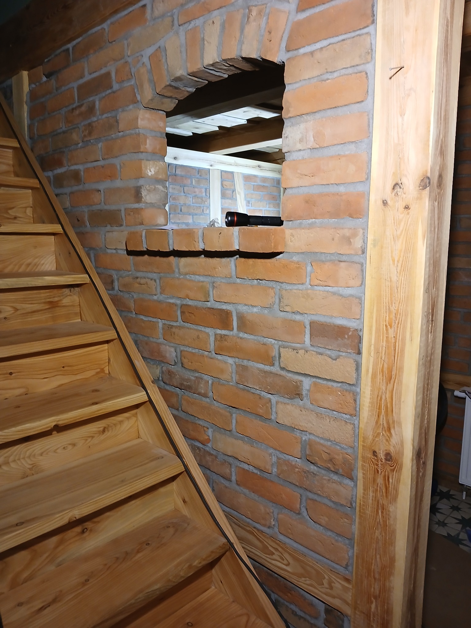 Murowana ściana z cegły z łukowym otworem, obok drewniane schody i słup z widocznym usłojeniem drewna, w otworze widoczna biała konstrukcja.