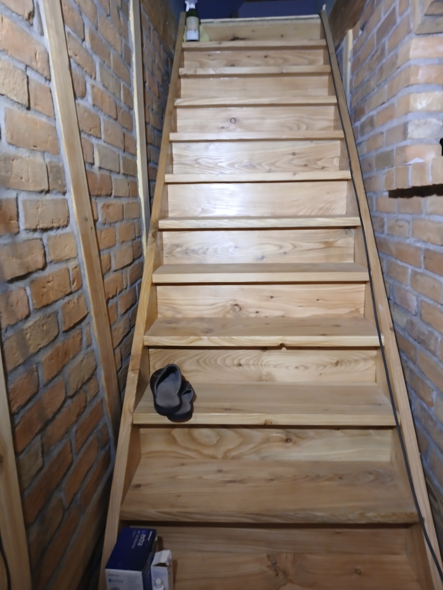 Drewniane schody z surowego drewna, prowadzące na górę, z ceglanymi ścianami po bokach, na schodach leżą klapki i pudełko.
