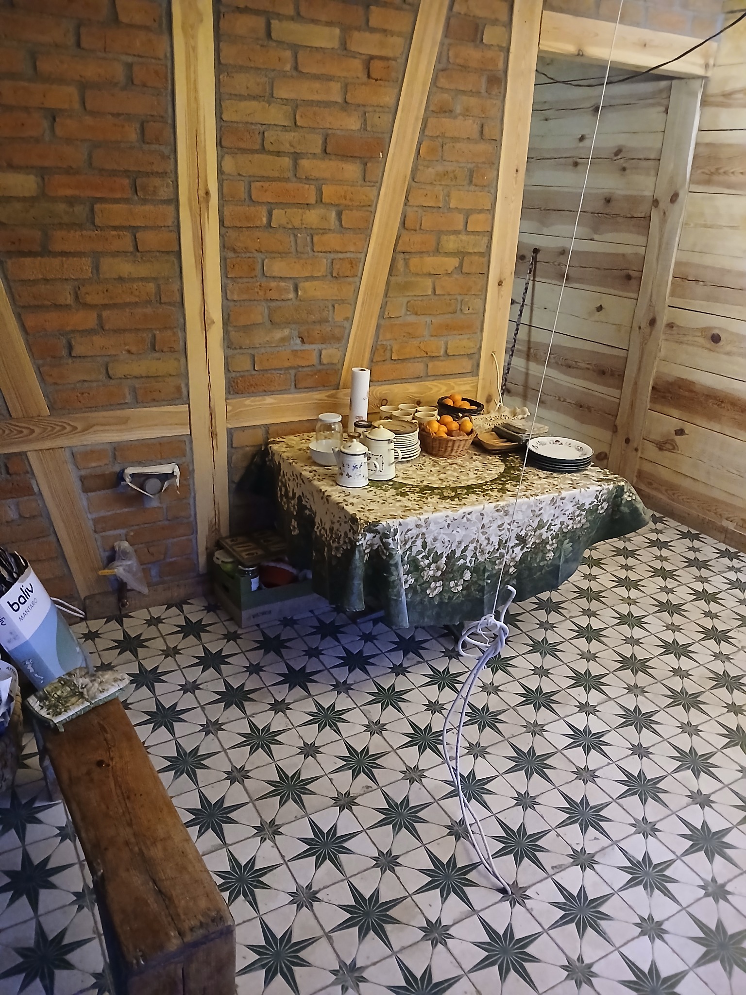 Wnętrze z ceglaną ścianą i drewnianą konstrukcją, stół nakryty kwiecistym obrusem z naczyniami i owocami, wzorzysta podłoga w gwiazdy.