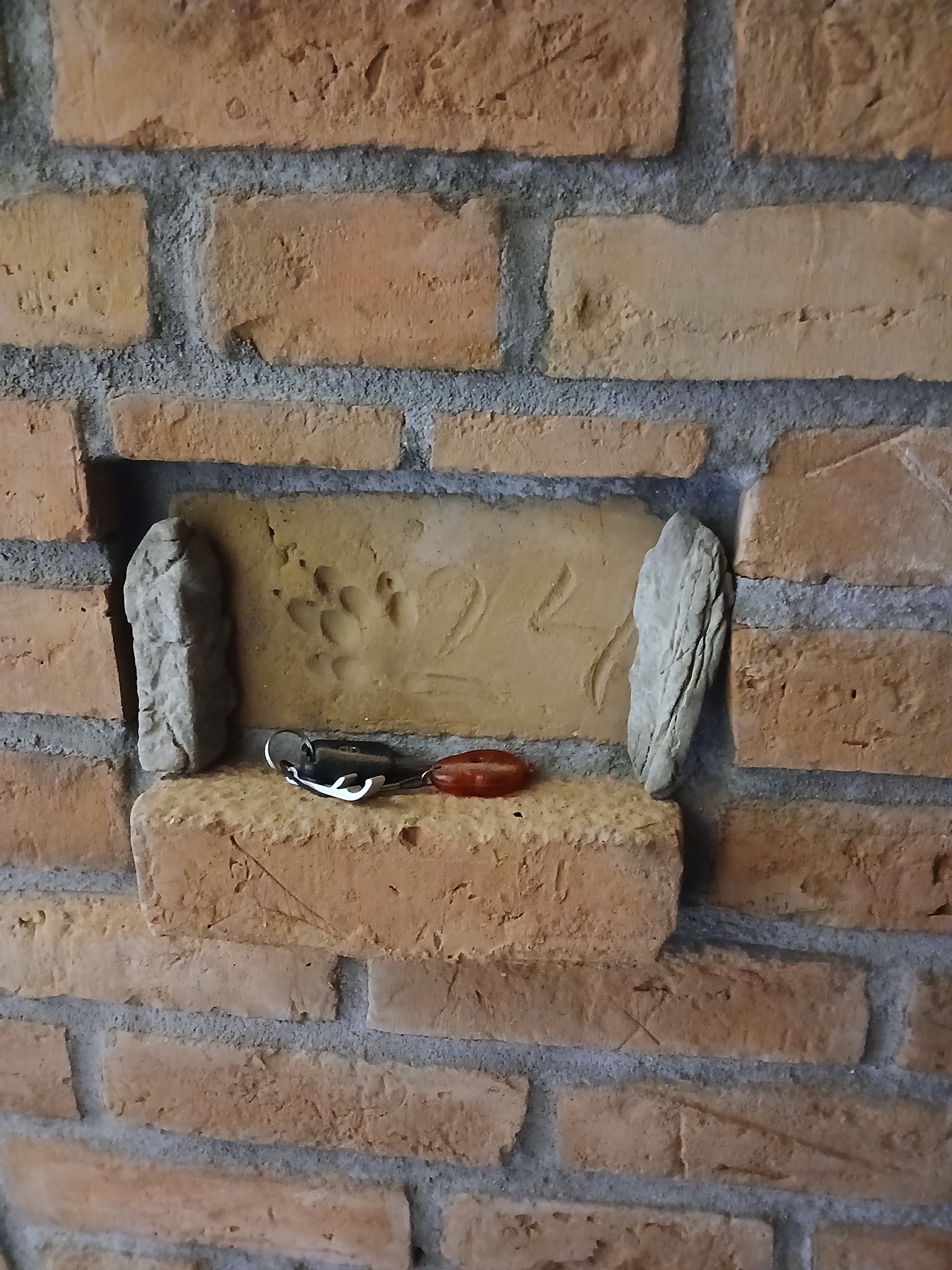 Murowana ściana z cegły z wnęką, w której znajdują się dwa kamienie, cegła z odciskiem łapy oraz klucze z brelokiem.