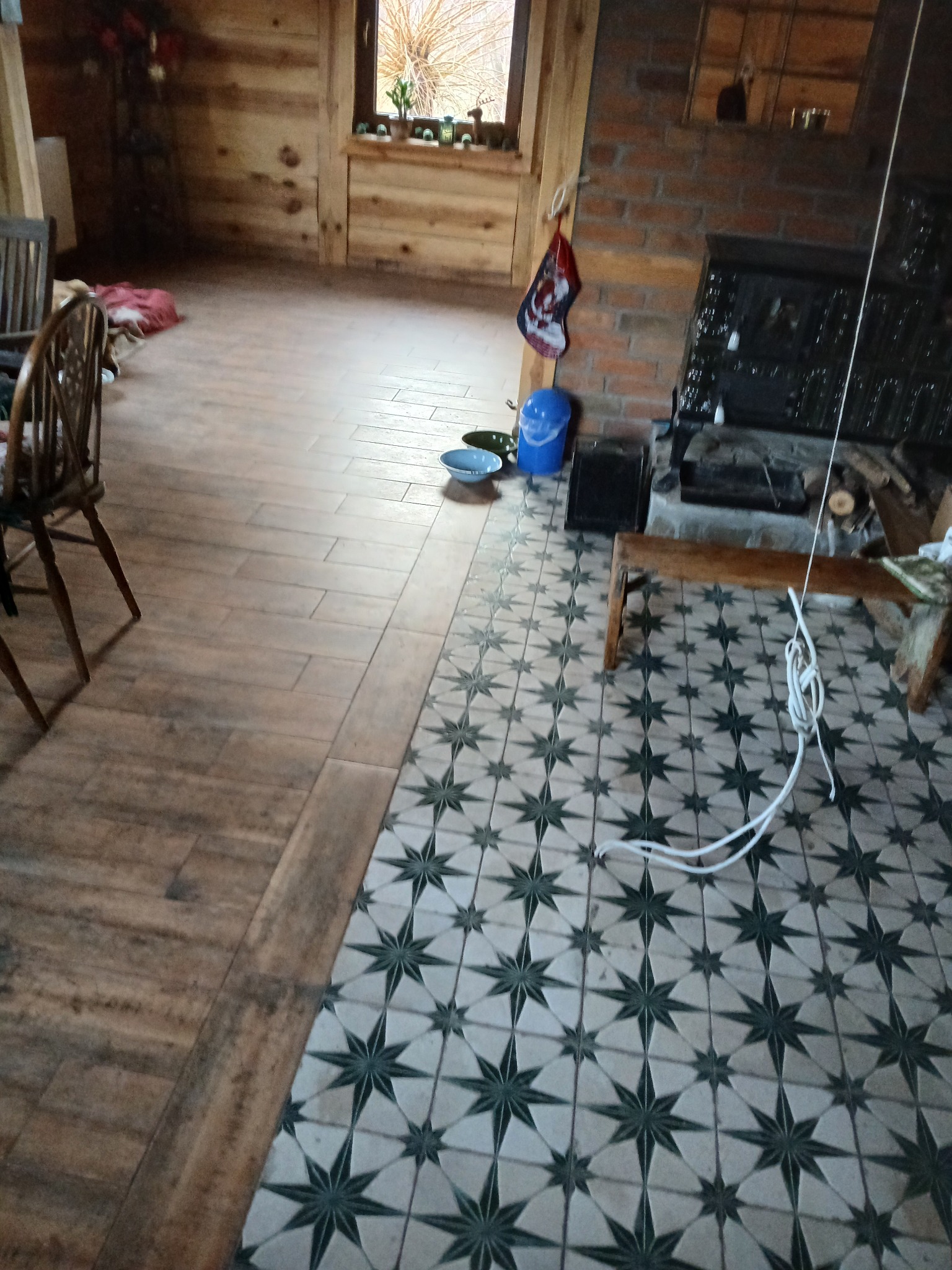 Połączenie dwóch rodzajów podłogi w drewnianym domu: ceramiczne płytki imitujące drewno i płytki w geometryczny wzór gwiazd, widoczny piec kaflowy i drewniane meble.