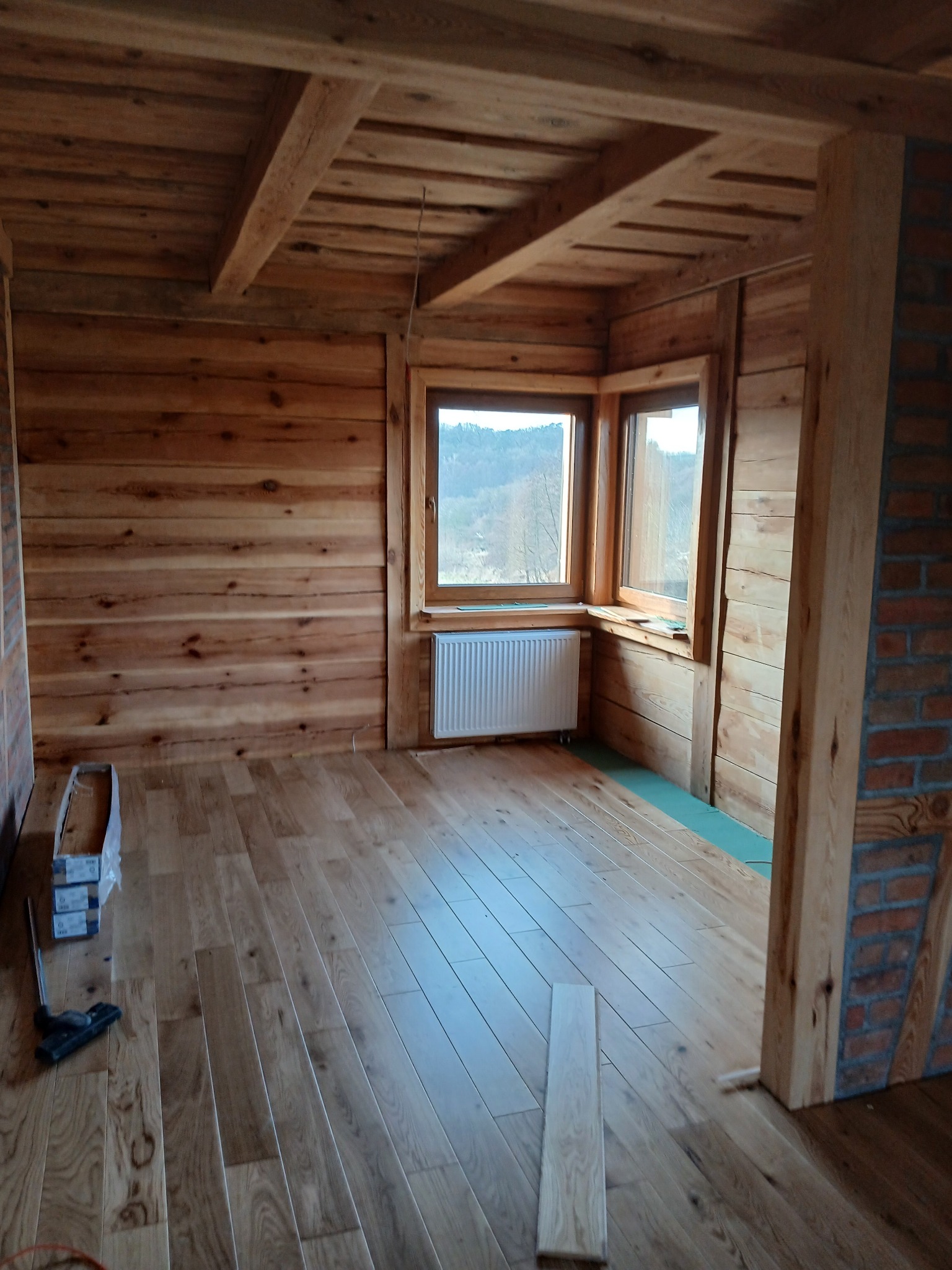 Wnętrze domu w Krępie z drewnianymi ścianami, podłogą i sufitem, widoczne okna, grzejnik oraz narzędzia i materiały do układania podłogi.