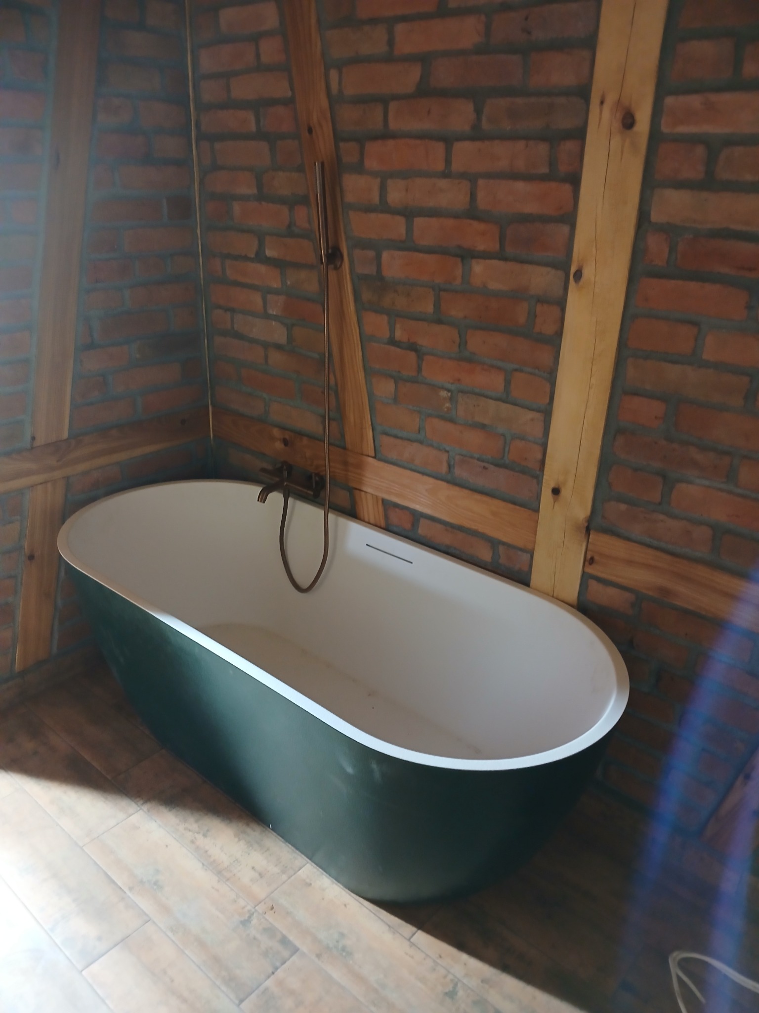 Wolnostojąca, owalna wanna w łazience z ceglanymi ścianami i drewnianą konstrukcją stropu, bateria w stylu retro.