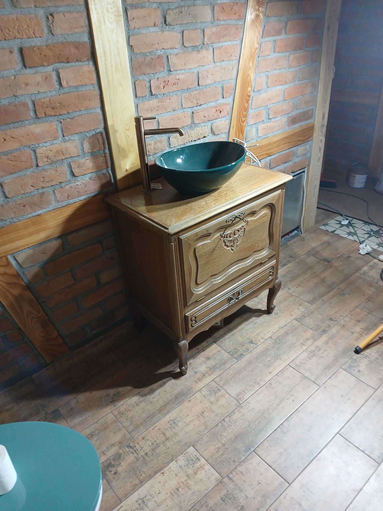 Umywalka wpuszczana w odrestaurowaną, drewnianą komodę z dekoracyjnymi rzeźbieniami, z nowoczesną, wysoką baterią, na tle ściany z cegły i drewnianych belek.