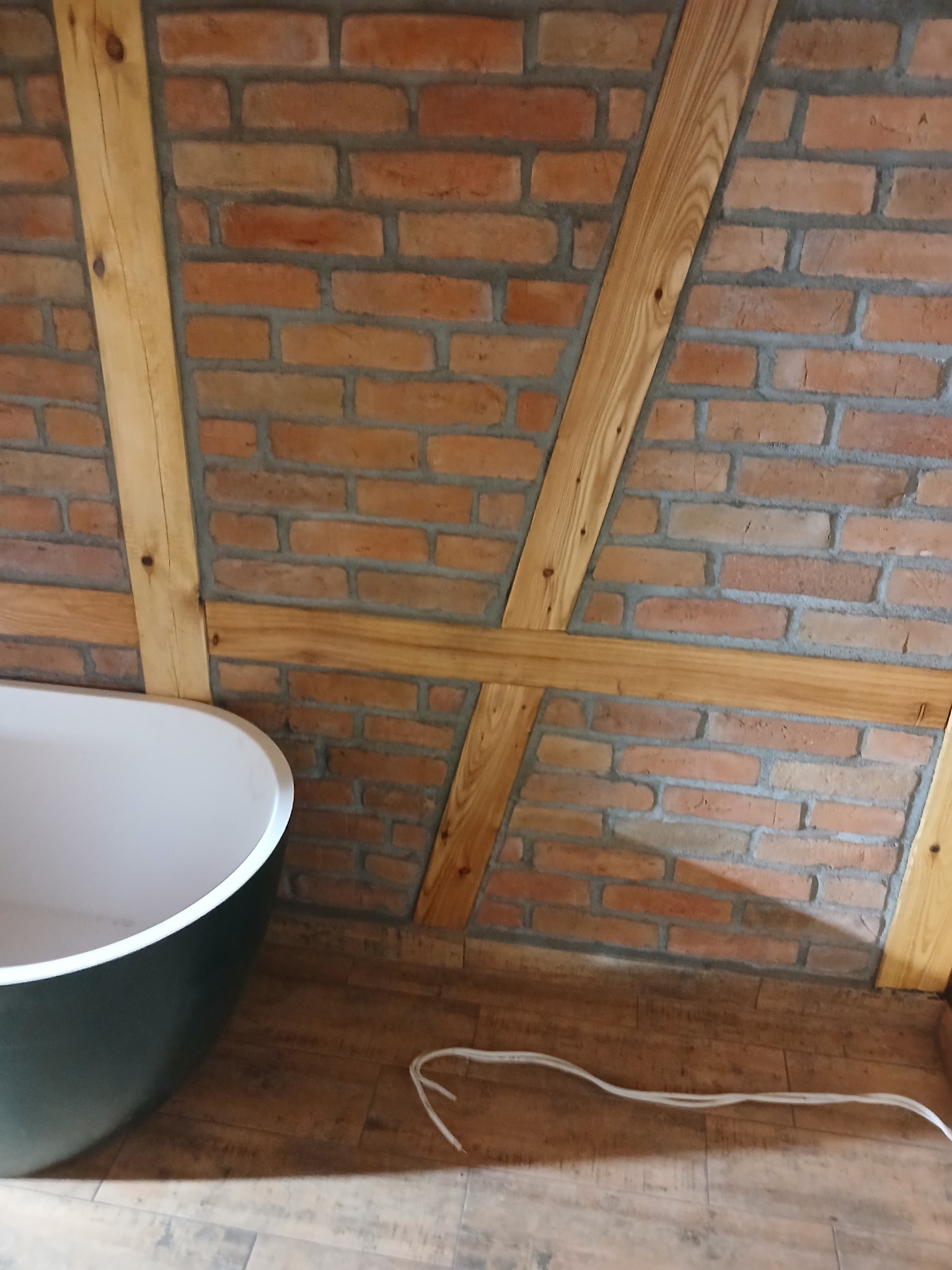 Ukośna ściana z czerwonej cegły z drewnianą konstrukcją i fragmentem ciemnej wanny w rogu, na podłodze leży kabel.