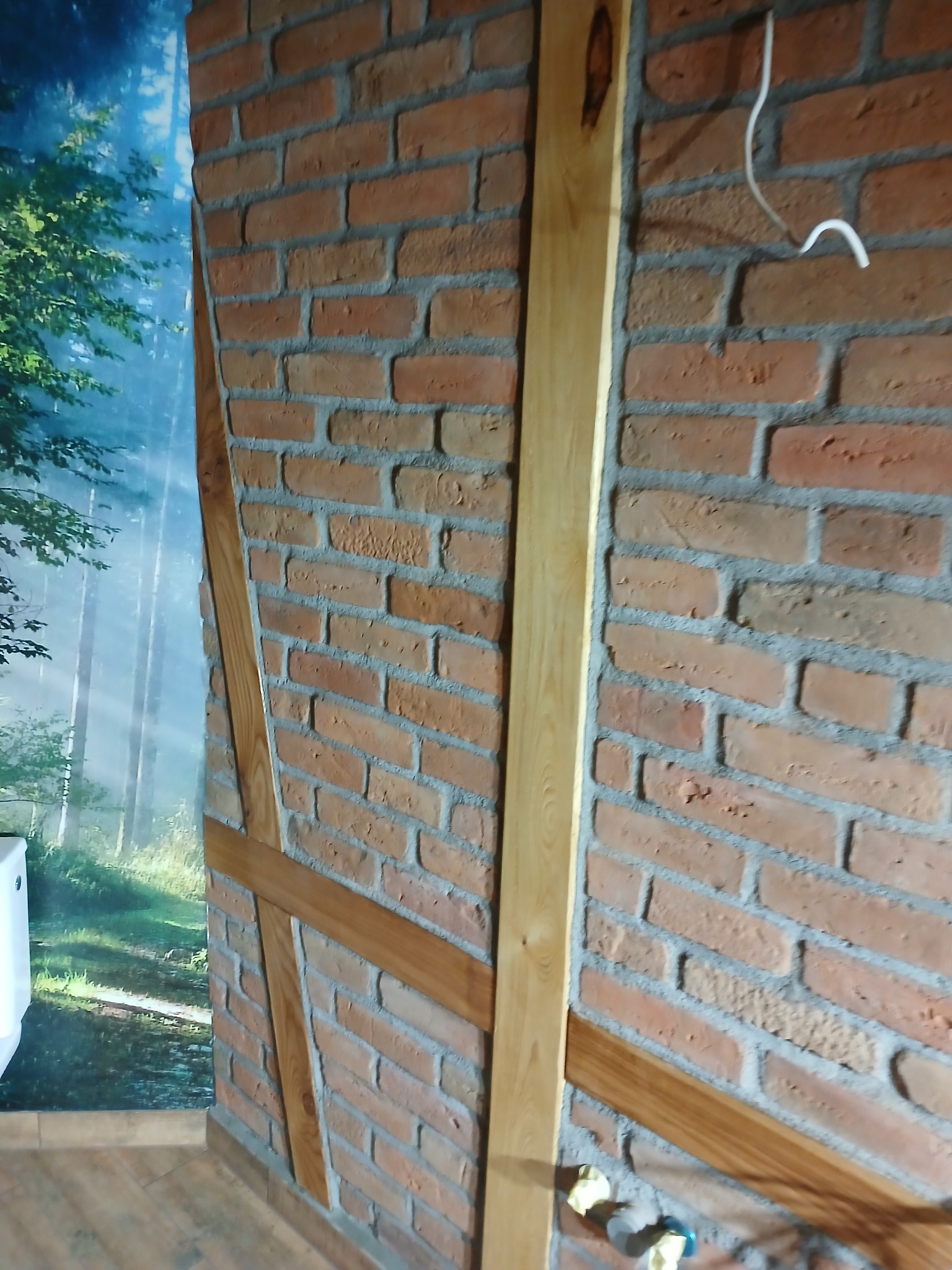 Ceglana ściana z drewnianymi elementami konstrukcyjnymi i tapetą z motywem leśnym w tle, widoczne przewody elektryczne i fragment instalacji wodnej.