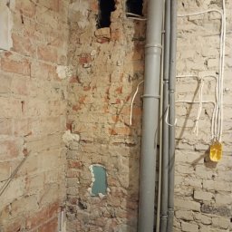 Piotr Rewaj - Odsłonięta cegła z instalacją hydrauliczną i elektryczną w trakcie remontu. Widoczne rury, kable, otwory w ścianie i pianka montażowa.