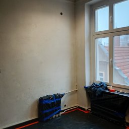 Piotr Rewaj - Pokój w trakcie remontu: ściany po szpachlowaniu, zabezpieczone podłogi folią, zakryty grzejnik. Widoczne otwory w ścianie i okno z widokiem na dach.