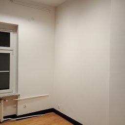 Piotr Rewaj - Pomalowane na biało wnętrze pokoju z drewnianą podłogą i czarnymi listwami przypodłogowymi. Widoczne okno, grzejnik i lampa sufitowa. Świeżo po remoncie.