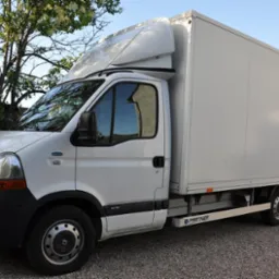 Biały samochód dostawczy typu furgon z nadbudówką, zaparkowany na żwirowym podłożu, z widocznym napisem 'PARTNER' na listwie progowej i odbiciem nieba w szybie.