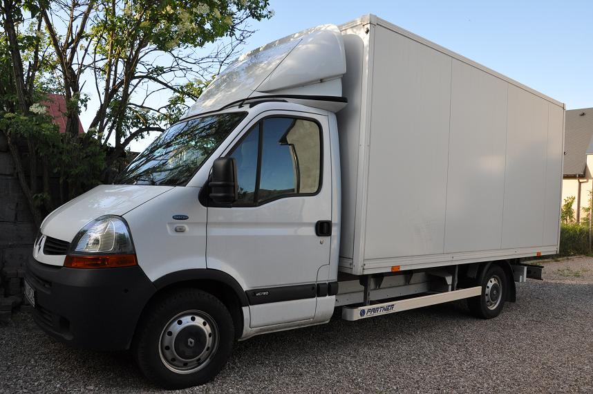 Biały samochód dostawczy typu furgon z nadbudówką, zaparkowany na żwirowym podłożu, z widocznym napisem 'PARTNER' na listwie progowej i odbiciem nieba w szybie.