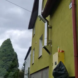 Zewnętrzny, stalowy komin spalinowy zamontowany na elewacji domu o oliwkowym odcieniu, z widocznymi elementami mocującymi i załamaniami rur.