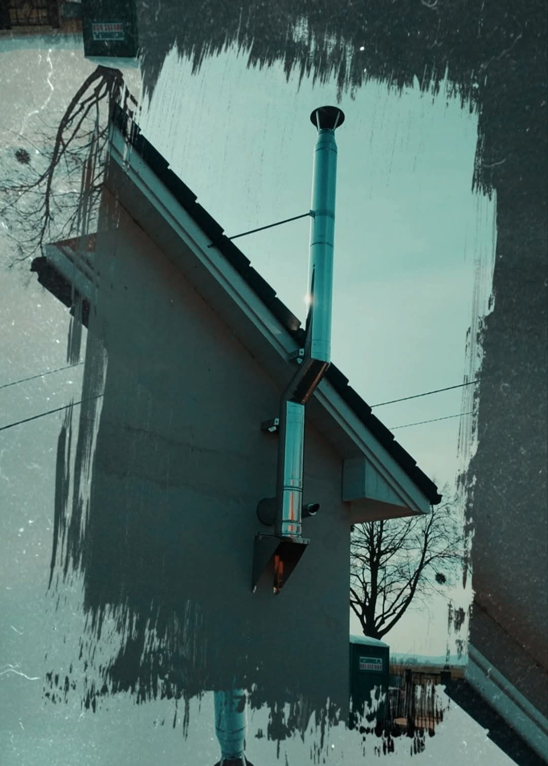 Zewnętrzny, metalowy komin systemowy, zamontowany przy ścianie domu z widocznym fragmentem dachu i drzewem w tle, z efektem postarzanej fotografii.