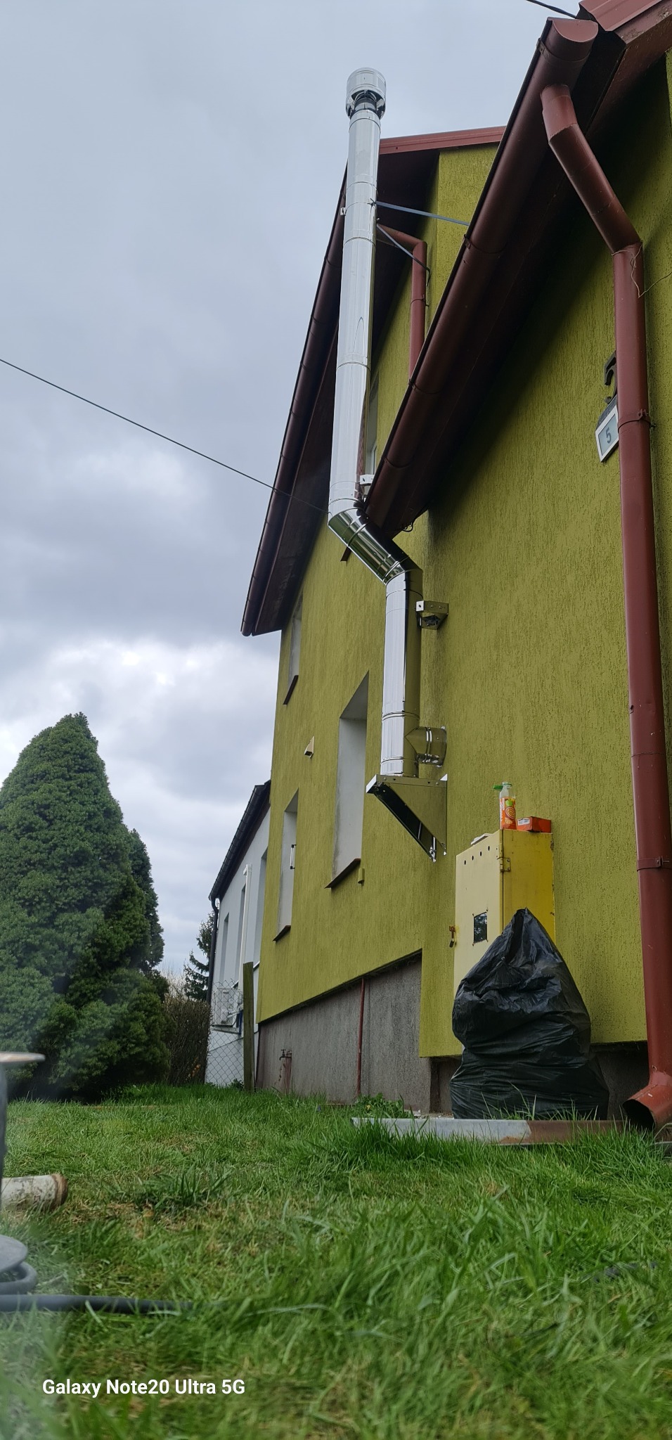 Zewnętrzny, stalowy komin spalinowy zamontowany na elewacji domu o oliwkowym odcieniu, z widocznymi elementami mocującymi i załamaniami rur.