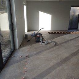 Dakota - Układanie płytek podłogowych w jasnym, nowoczesnym wnętrzu. Widoczne krzyżyki dystansowe pomiędzy płytkami, odkurzacz przemysłowy, drabina i materiały budowlane.