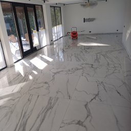 Dakota - Salon z nową, błyszczącą podłogą z płytek imitujących marmur, widoczne duże przesuwne drzwi balkonowe i odkurzacz przemysłowy w rogu pomieszczenia.