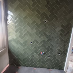 Dakota - Ściana wyłożona zielonymi, błyszczącymi płytkami ułożonymi w jodełkę, z widocznymi otworami na instalacje i plastikowymi krzyżykami dystansowymi na podłodze.