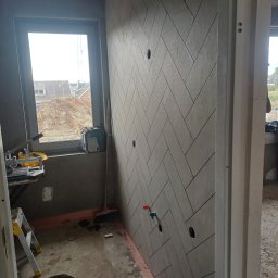 Dakota - Ściana wyłożona szarymi płytkami w jodełkę, widoczne otwory na instalacje, obok okno z widokiem na plac budowy, w tle narzędzia i drabina.