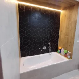 Nowoczesna łazienka z wanną obudowaną płytkami w odcieniach szarości, czarna ściana z geometrycznym wzorem płytek i drewnopodobna zabudowa z oświetleniem LED