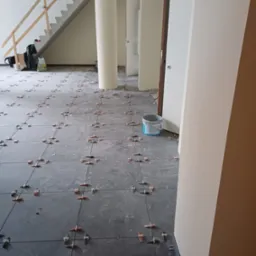 Układanie szarych płytek podłogowych w przestronnym wnętrzu, z widocznymi krzyżykami dystansowymi i wiadrem z zaprawą, w tle schody i kolumny.