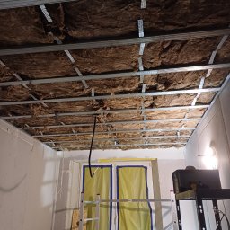 Dakota - Montaż izolacji z wełny mineralnej w suficie podwieszanym z metalowym stelażem, widoczne profile CD i UD, oraz fragment ściany z płyt kartonowo-gipsowych i drabina.