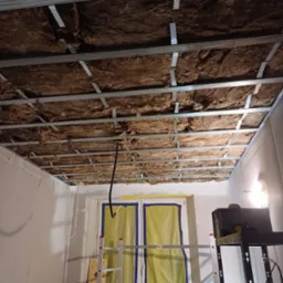 Montaż izolacji z wełny mineralnej w suficie podwieszanym z metalowym stelażem, widoczne profile CD i UD, oraz fragment ściany z płyt kartonowo-gipsowych i drabina.