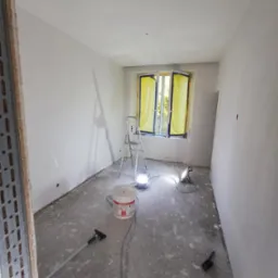 Pokój w trakcie prac wykończeniowych: świeżo wyszpachlowane ściany, zabezpieczone folią okno, drabina, lampa budowlana oświetlająca wiadro z masą szpachlową Unifix S3 i narzędzia na zaszpachlowanej...