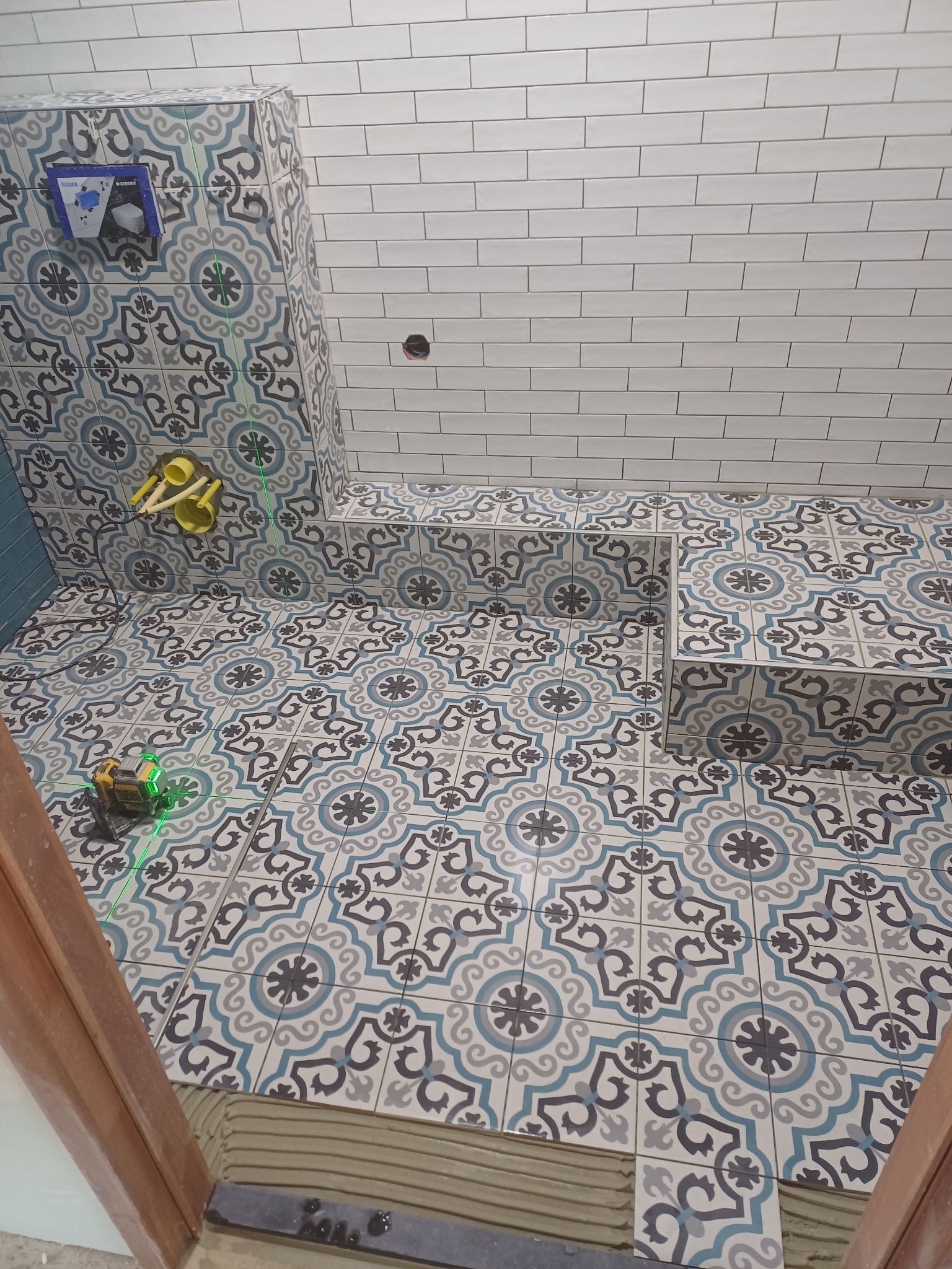 Łazienka w trakcie wykańczania: wzorzyste płytki podłogowe i ścienne w odcieniach błękitu i szarości, białe, prostokątne płytki na ścianie, poziomica laserowa ustawiona na podłodze, widoczne...