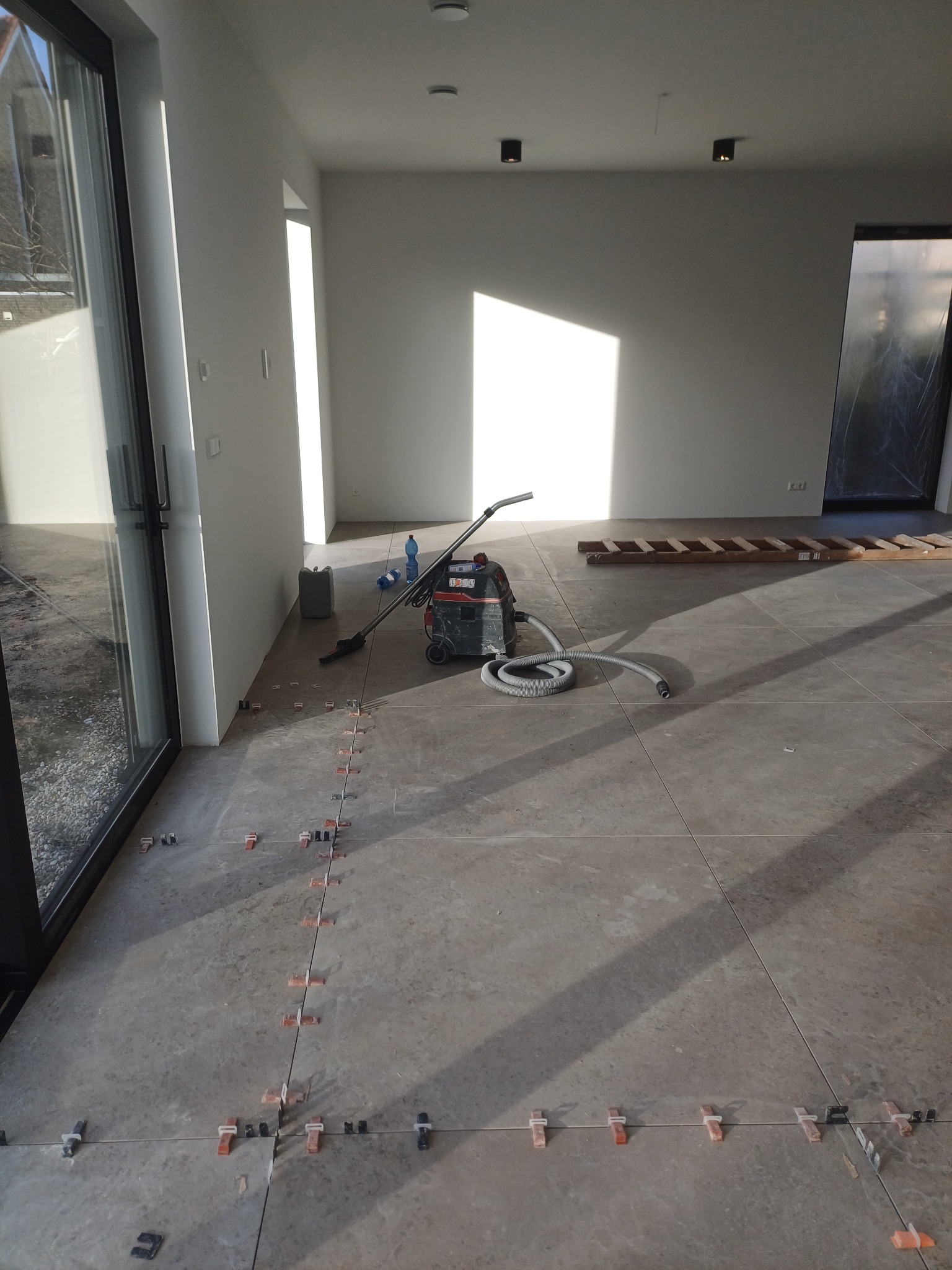 Układanie płytek podłogowych w jasnym, nowoczesnym wnętrzu. Widoczne krzyżyki dystansowe pomiędzy płytkami, odkurzacz przemysłowy, drabina i materiały budowlane.