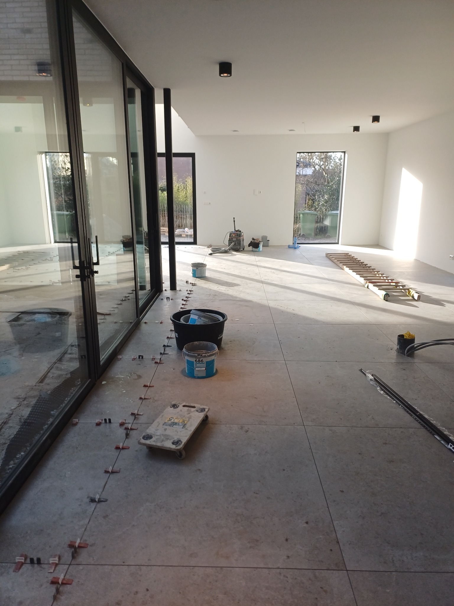 Układanie płytek podłogowych w przestronnym, jasnym wnętrzu z dużymi oknami; widoczne poziomice, wiadra z zaprawą, krzyżyki dystansowe i wózek do transportu płytek.