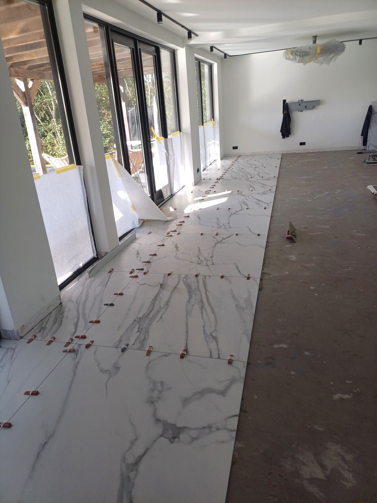 Układanie marmurowych płytek podłogowych w jasnym pomieszczeniu z dużymi oknami, widoczne krzyżyki dystansowe i część niewykończonej podłogi.