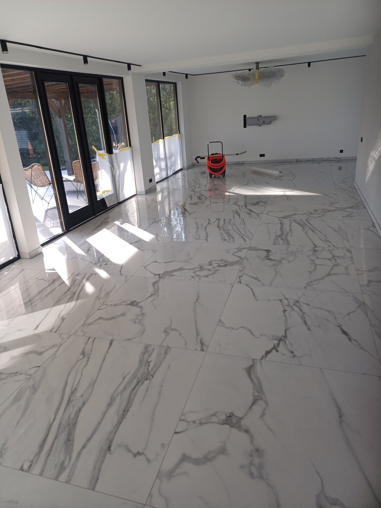 Salon z nową, błyszczącą podłogą z płytek imitujących marmur, widoczne duże przesuwne drzwi balkonowe i odkurzacz przemysłowy w rogu pomieszczenia.