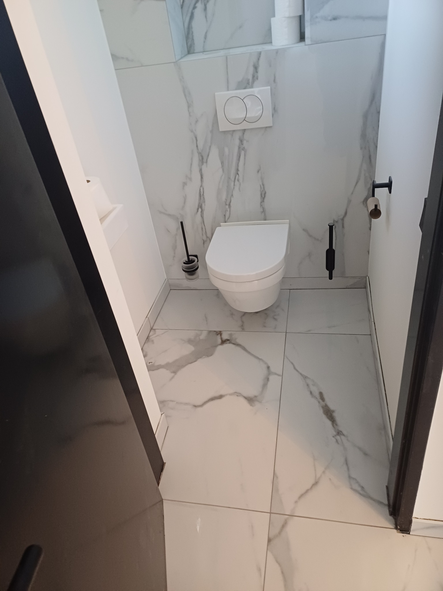 Nowoczesna toaleta podwieszana w łazience wykończonej białymi płytkami imitującymi marmur, z czarnymi akcentami w postaci szczotki toaletowej i uchwytu na papier, widok zza uchylonych drzwi.