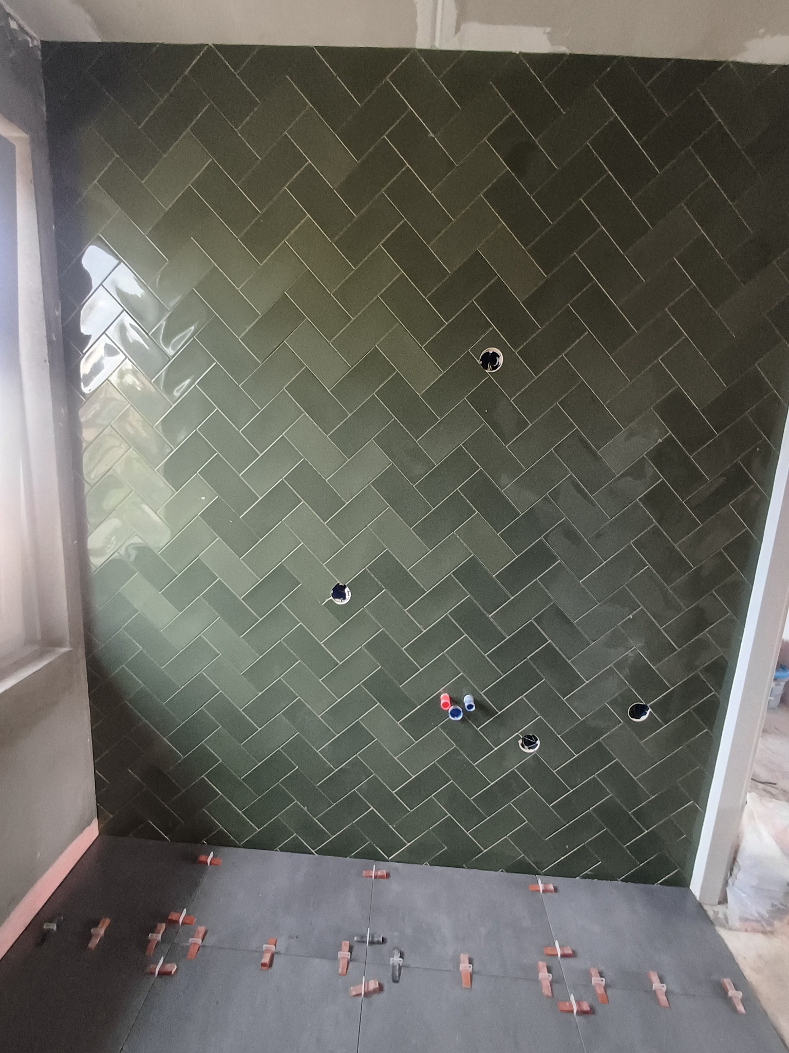 Ściana wyłożona zielonymi, błyszczącymi płytkami ułożonymi w jodełkę, z widocznymi otworami na instalacje i plastikowymi krzyżykami dystansowymi na podłodze.