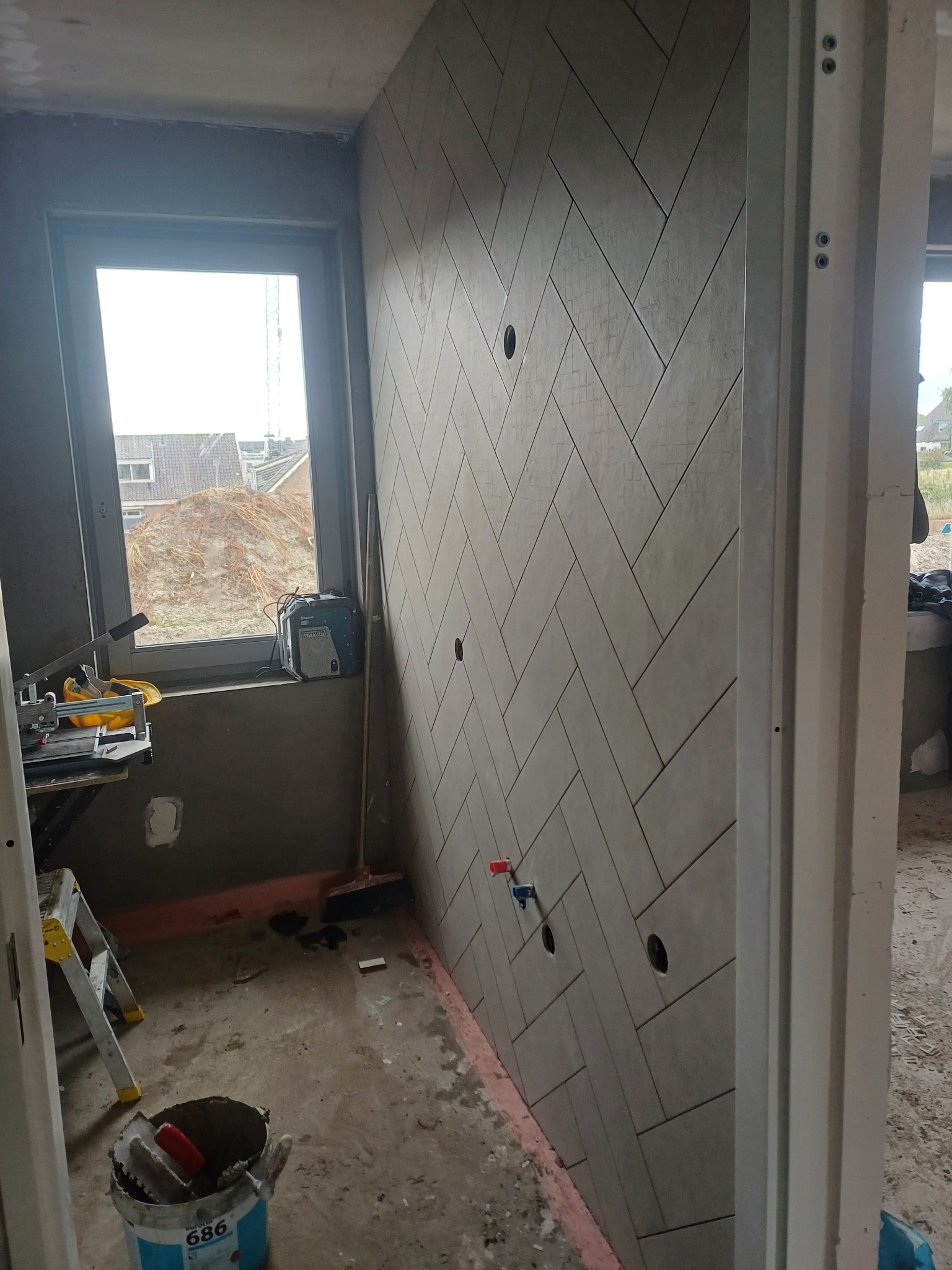 Ściana wyłożona szarymi płytkami w jodełkę, widoczne otwory na instalacje, obok okno z widokiem na plac budowy, w tle narzędzia i drabina.